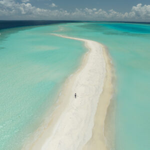 A high drone shot of long sandbank strip of white sand with a lady walking out into the distance, surrounded by cerulean blue water either side.