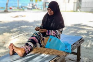 A lady on Kamadhoo, Maldives sitting performing local craftwork, with straw, on a beautiful island in the shade on a sunny day.