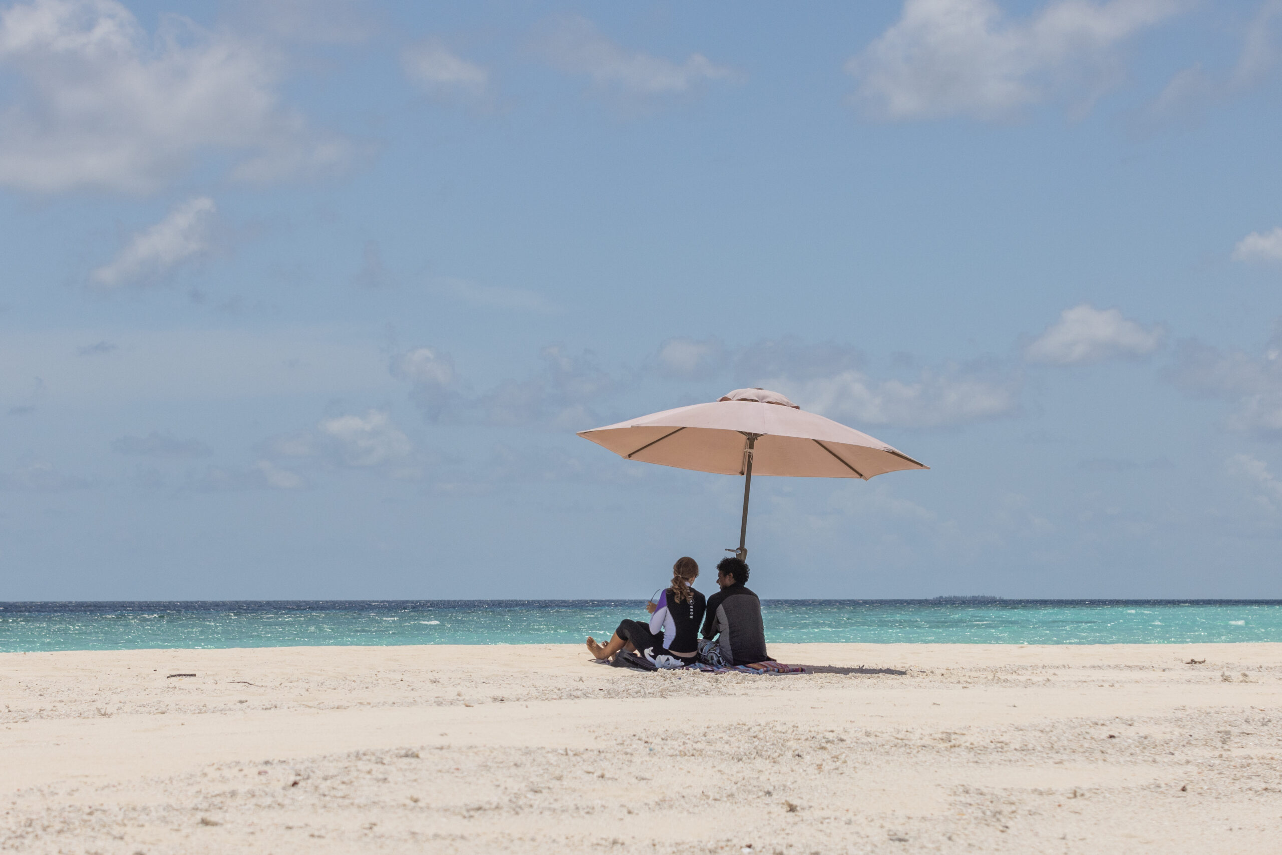 Two people on a white sandy Maldivian beach, under an umbrella in the sun.